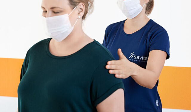 Die junge Trainerin steht hinter der Patientin. Die Patientin hat die Augen geschlossen. Die Trainerin schlägt vorsichtig auf die linke Schulter der Patientin. Beide tragen einen Mund-Nasen-Schutz.
