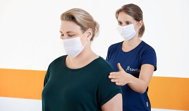 Die junge Trainerin steht hinter der Patientin. Die Patientin hat die Augen geschlossen. Die Trainerin schlägt vorsichtig auf die linke Schulter der Patientin. Beide tragen einen Mund-Nasen-Schutz.