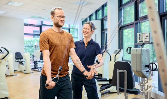 Ein Mann trainiert an einem Seilzuggerät im Therapie- und Trainingszentrum Neuss, während eine Trainerin ihn bei der Übung begleitet.