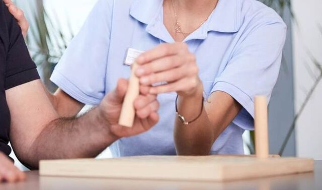 Die attraktive Ergotherapeutin unterstützt den neurologischen Patienten bei der Ausführung einer Greifübung. Beide lächeln.