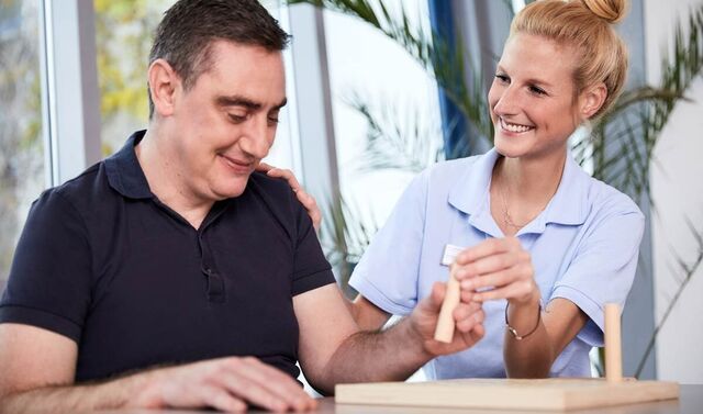 Die attraktive Ergotherapeutin unterstützt den neurologischen Patienten bei der Ausführung einer Greifübung. Beide lächeln.