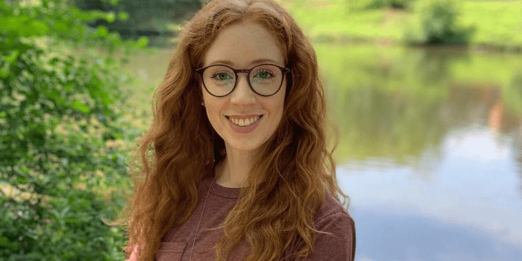 Marie Erkes steht in der Landschaft vor einem Teich und lächelt in die Kamera.