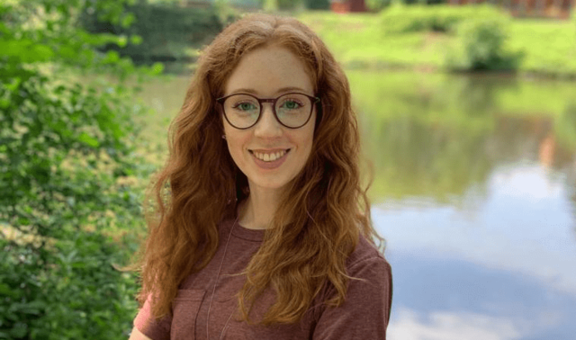 Marie Erkes steht in der Landschaft vor einem Teich und lächelt in die Kamera.