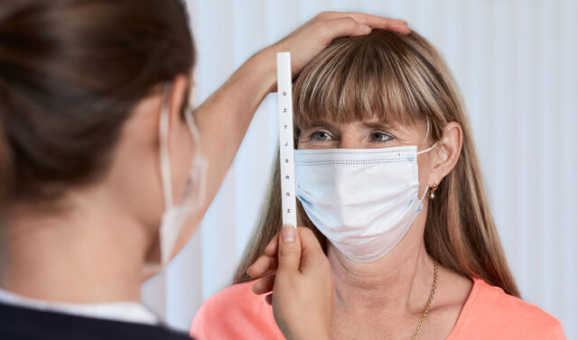 Eine Frau mit Mund-Nasen-Schutz schaut auf einen Stab mit Buchstaben. Im Vordergrund ist der Hinterkopf der Trainerin zu sehen, die den Stab hält und eine Hand auf den Kopf der Patientin legt.