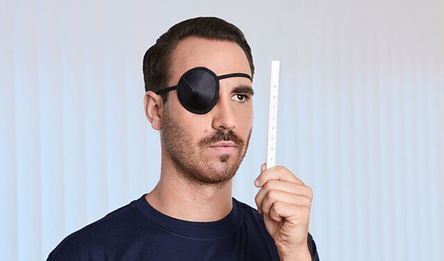 Ein junger, attraktiver Mann trägt eine Augenklappe und schaut mit dem offenen Auge auf einen Stab mit Buchstaben.