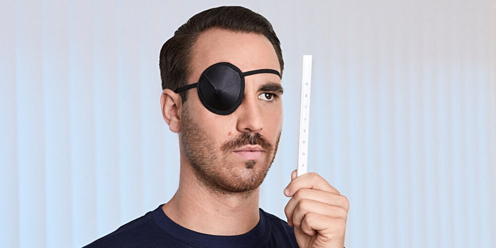 Ein junger, attraktiver Mann trägt eine Augenklappe und schaut mit dem offenen Auge auf einen Stab mit Buchstaben.