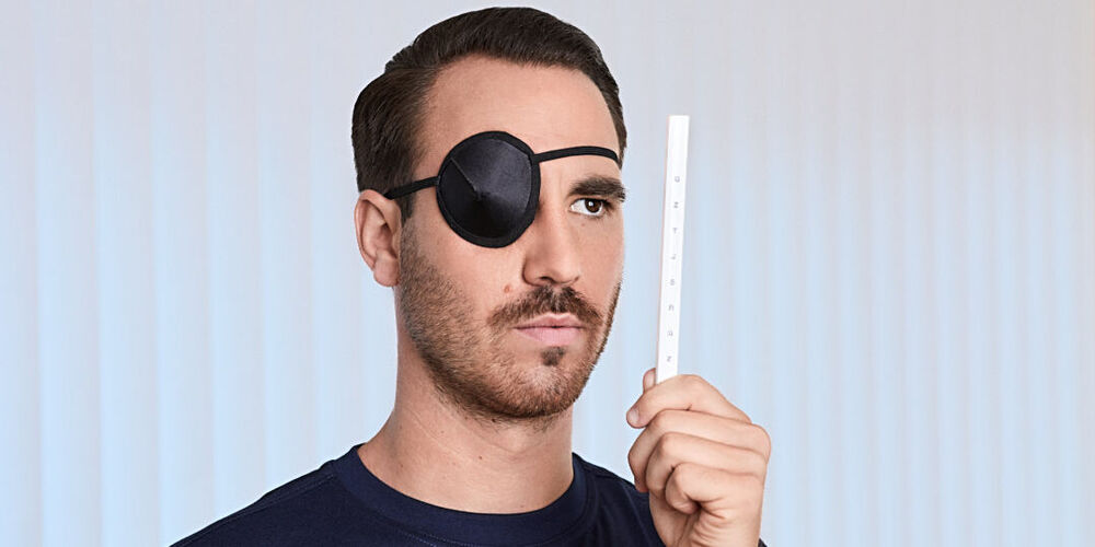 Ein junger, attraktiver Mann trägt eine Augenklappe und schaut mit dem offenen Auge auf einen Stab mit Buchstaben.