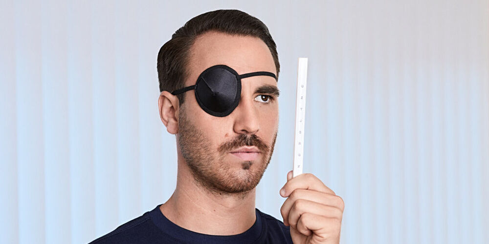 Ein junger, attraktiver Mann trägt eine Augenklappe und schaut mit dem offenen Auge auf einen Stab mit Buchstaben.