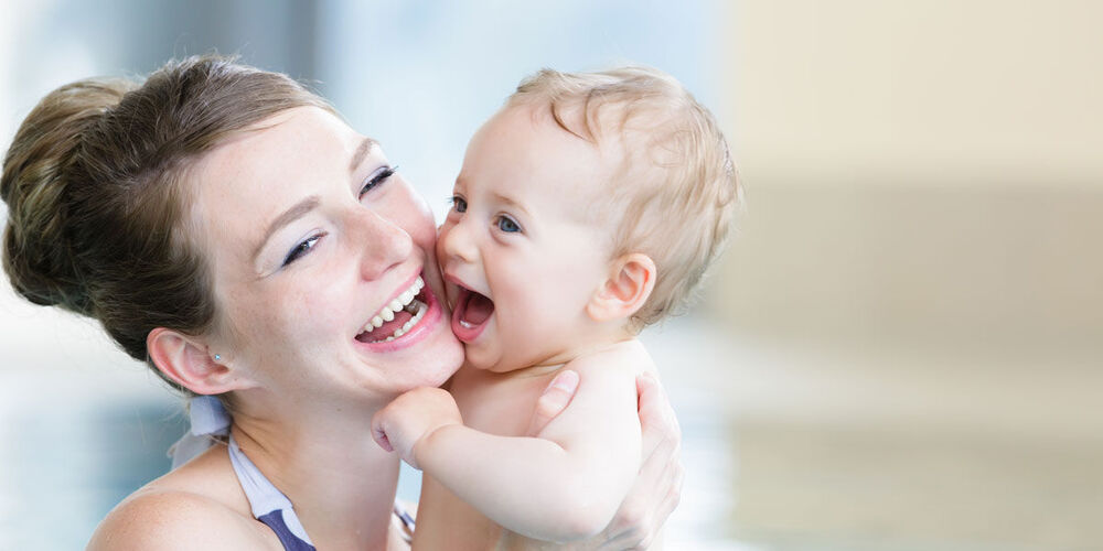 Eine Mutter mit ihrem Baby in einem Schwimmbecken. Beide lachen und die Mutter hält ihr Kind an ihre Wange gedrückt. 