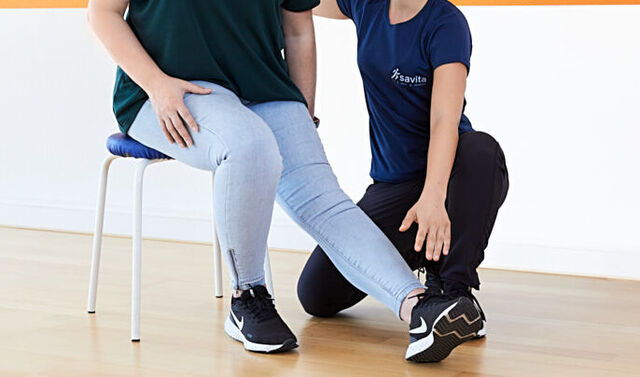 Therapiesituation aus dem Neurozentrierten Training. Die Trainerin führt mit der Patientin eine Nervendehnung durch. Dazu sitzt die Patientin auf einem Stuhl. Ihr linkes Bein ist gestreckt und der gestreckte Fuß nach innen gedreht. Beide Personen tragen einen Mund-Nasen-Schutz.