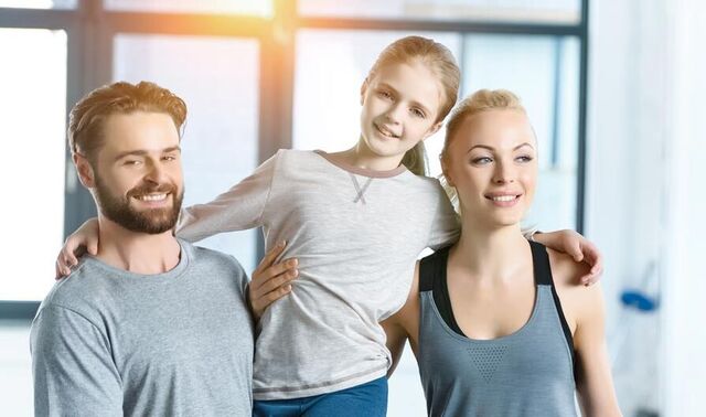 Eine Familie bestehend aus Eltern und junger Tochter stehen in Sportkleidung in einem Fitnessraum. Der Papa hält die Tochter auf dem Arm.