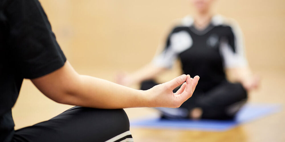 Im Vordergrund sieht man den Rücken und die recht Hand von einer Person, die im Schneidersitzt sitzt und den Arm offen auf dem Bein abgelegt hat. Im Hintergrund sitzt eine Person in der gleichen Meditationspose.