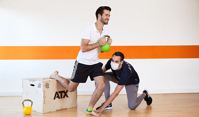 Ein junger Therapeut führt eine Trainingsübung mit einem jungen Sportler durch. Der Fuß ist durch ein Kinesiotape getaped. Der Sportler hält ein grünes Gewicht in der Hand. Das hintere Bein liegt angewinkelt auf einem Kasten. Das vordere Bein macht eine Kniebeuge. Der Therapeut trägt eine Maske und kniet neben dem Sportler und stabilisiert mit den Händen Knie und Fuß.