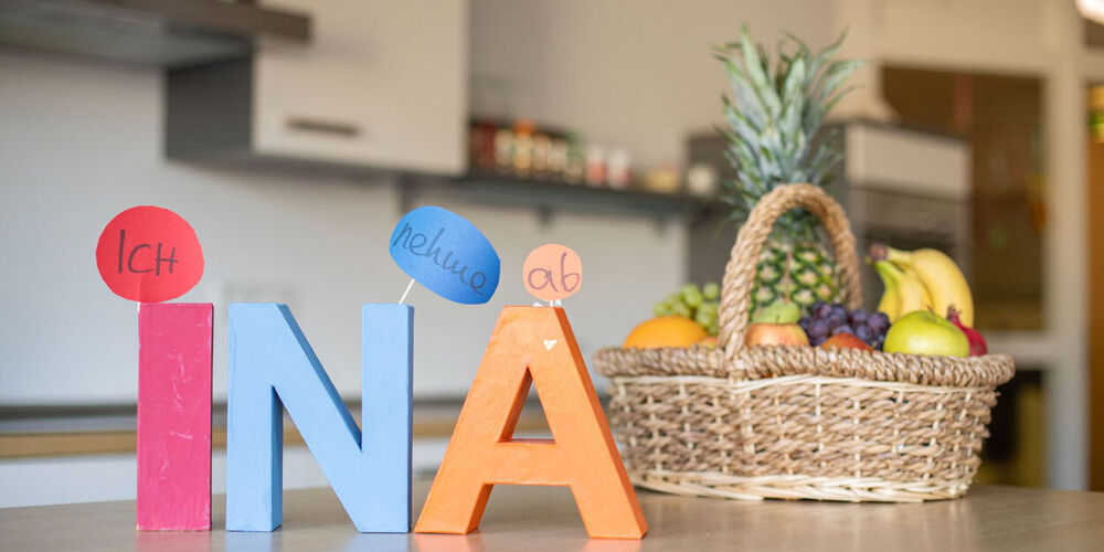 Im Vordergrund stehen die bunten Holzbuchstaben "INA". Leicht dahinter ist ein gefüllter Obstkorb zu sehen. Im Hintergrund ist die Küche zu sehen.