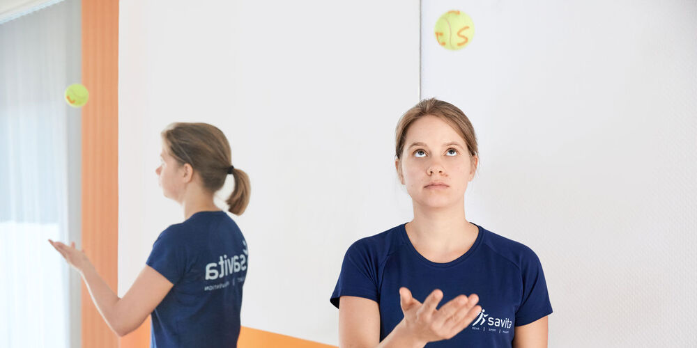 Ein junge Trainerin in blauem savita Shirt wirft einen Tennisball mit Buchstaben hoch. Sie steht vor einem Spiegel, der die Aktion gespiegelt abbildet.