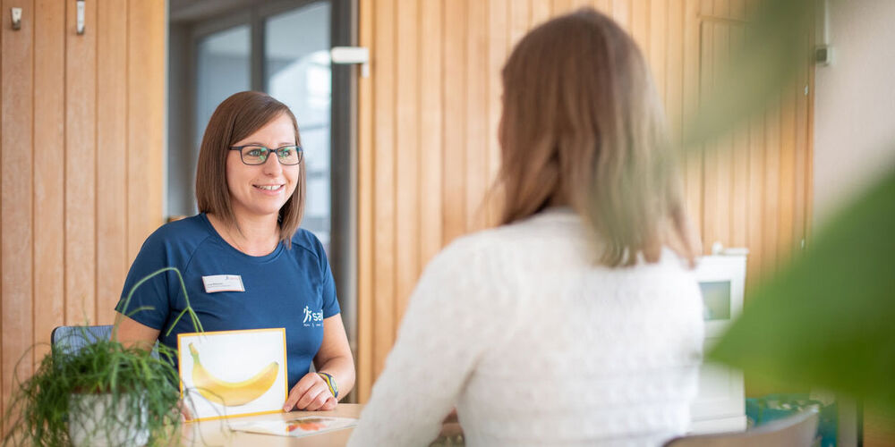 Logopädin zeigt der Patientin eine Karte mit einer abgebildeten Banane und übt mit ihr die Aussprache.