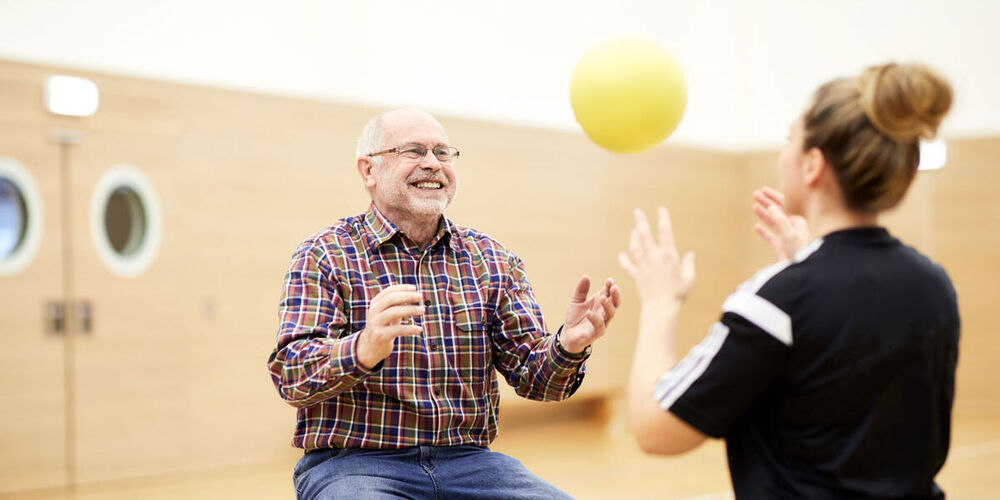 Eine Therapeutin wirft einem sitzenden älteren Patienten einen gelben Ball zu. Dieser fängt ihn gut gelaunt.