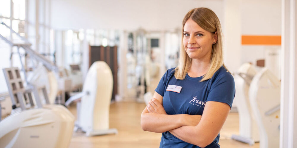 Attraktive Therapeutin steht mit verschränkten Armen vor der hellen Trainingsfläche und schaut freundlich in die Kamera.
