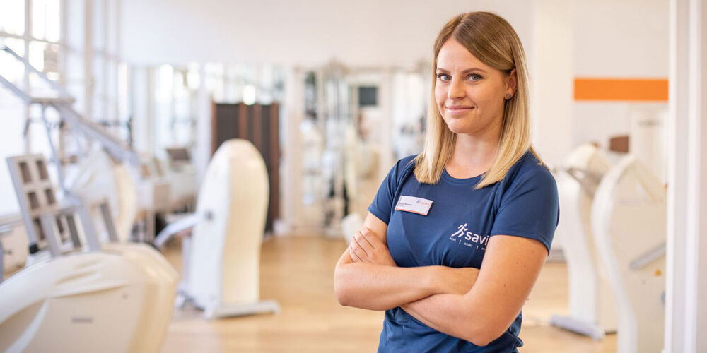 Attraktive Therapeutin steht mit verschränkten Armen vor der hellen Trainingsfläche und schaut freundlich in die Kamera.