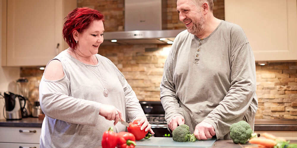 Eine Frau und ein Mann stehen in der Küche und schnippeln Gemüse. Beide lächeln.