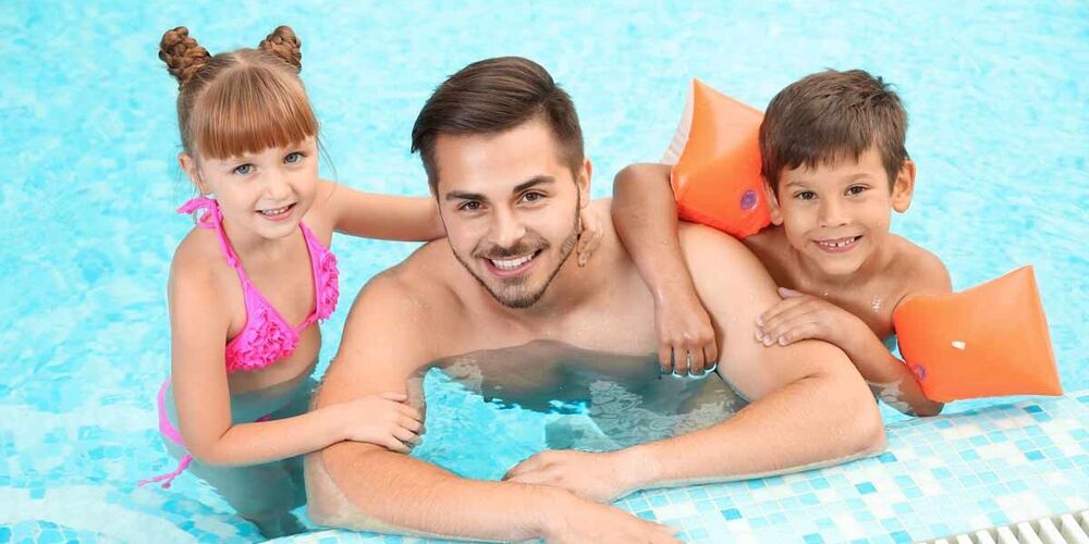 Ein junger Mann ist mit einem kleinen Jungen, welcher Schwimmflügel trägt und einem kleinem Mädchen im Schwimmbad am Beckenrad. Sie schauen alle drei lächelnd in die Kamera und halten sich am Beckenrad fest. 
