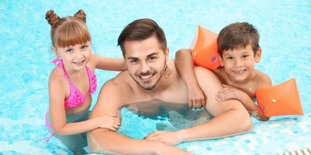 Ein junger Mann ist mit einem kleinen Jungen, welcher Schwimmflügel trägt und einem kleinem Mädchen im Schwimmbad am Beckenrad. Sie schauen alle drei lächelnd in die Kamera und halten sich am Beckenrad fest.