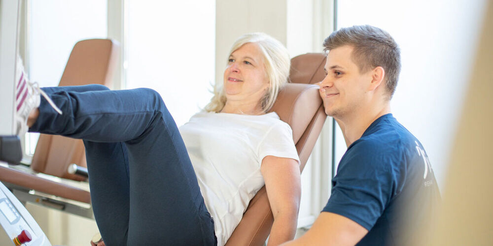 Patientin führt unter Anleitung eines Trainers eine Übung an der Beinpresse durch.