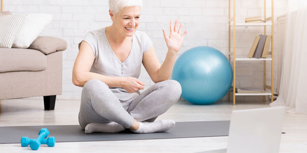 Ältere Damen in Sportkleidung sitzt auf einer Trainingsmatte und winkt in ihren Laptop.