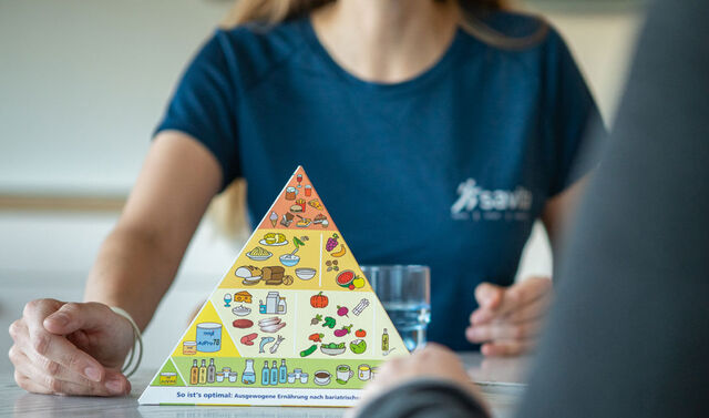 Im Vordergrund ist eine Ernährungspyramide zu sehen. Im Hintergrund befindet sich die Ernährungsberaterin im Gespräch mit einem Patienten.
