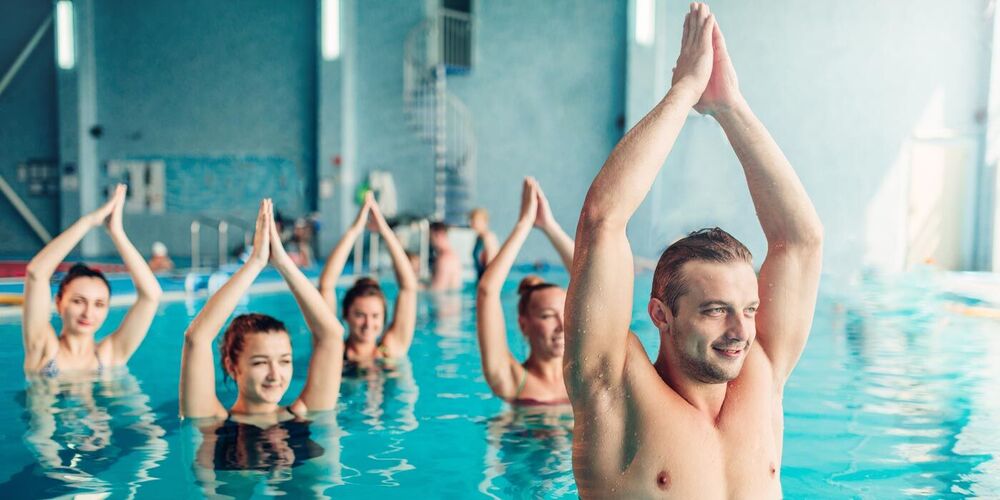 Eine Gruppe Erwachsener stehen in einem Schwimmbad im Wasser. Die Hände fassen über dem kopf zusammen. Im Vordergrund ist ein attraktiver junger Mann zu sehen.