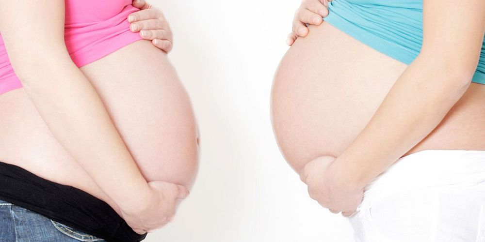 Zwei Schwangere stehen sich mit ihren Babybäuchen gegenüber. Beide halten die Bäuche mit ihren Händen.