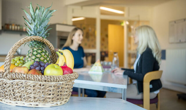 Im Vordergrund ist ein großer Obstkorb mit Bananen, Äpfeln, einer Ananas und einigem mehr. Im Hintergrund befinden sich zwei Frauen, die sich gegenüber an einem Tisch sitzen.