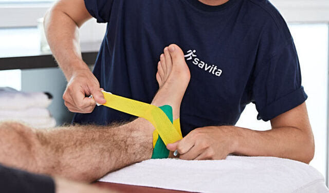Ein junger Therapeut klebt ein Kinesiotape auf den Fuß eines Mannes. Er trägt einen Mund-Nasen-Schutz und schaut nach vorne.